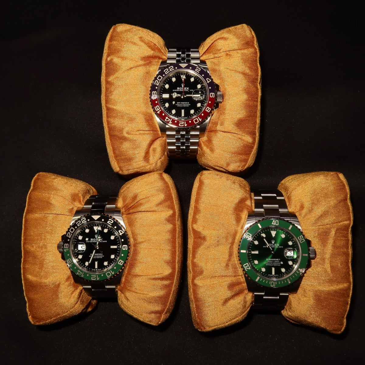 Explore luxury wristwatches: three Rolex sports watches with colored bezels on gold cushions.